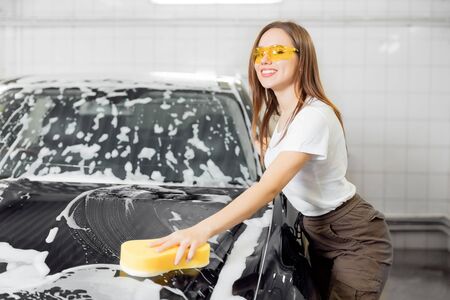 Car washing service. Worker beautiful woman cleaning auto black foam with yellow spongeの写真素材