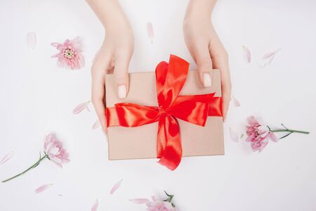 Hands of girl holding present gift craft box with red bow on white background with flowers, flat lay. Concept mother day, valentine holiday.の写真素材