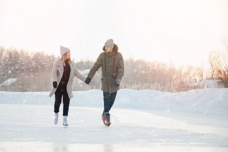 Girl and man are holding hands looking each other and eating on frozen ice of lake on skates. In background is sunsetの写真素材