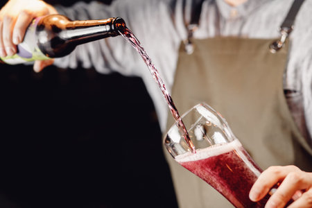 Barman pours craft blueberry cherry beer from bottle into glass, dark background.
