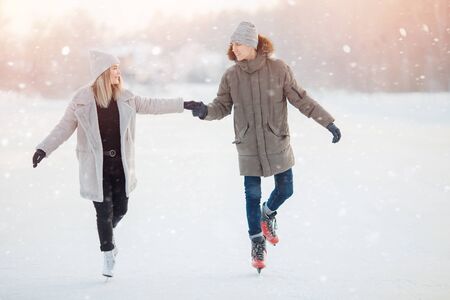 Loving couple in winter with skates on rink on sunset background, guy and girl dressed in warm hatの写真素材