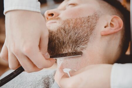 Hairdresser barber is holding razor for beard, male client is sitting in chair.の写真素材