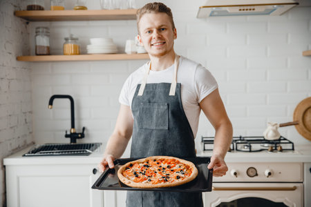 European male chef smiles and holds baking tray with hot Italian pizza in bright kitchen