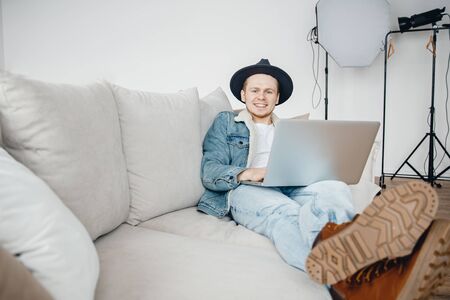 Freelancer photographer in hat video blogger works laptop, mounts videos for vlog about travel, in background lighting equipmentの写真素材