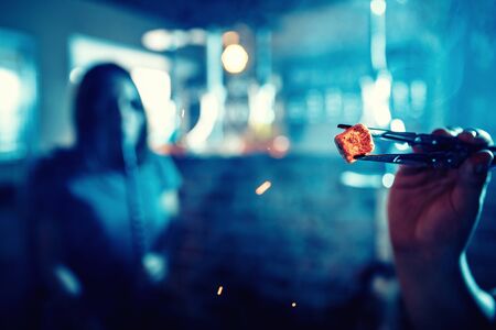 Hookah man changes and inflates hot coals for shisha tobacco tweezers red sparks, blue backgroundの写真素材