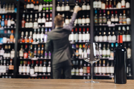 Bottle red wine and glass goblet on wooden table, in background sommelier chooses drink from...