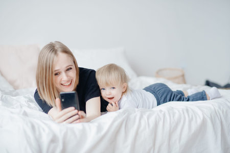 Young woman mom makes selfie photo with baby in bed on cell phone. Concept online chattingの写真素材