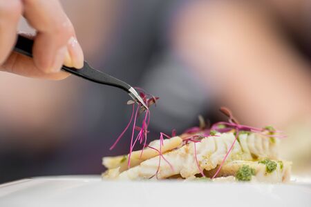 Chef with tweezers decorates with microgreens sprouts fish dish. Concept high art cooking, food decoration.