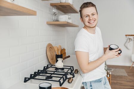 Young man smiles and shares freshly prepared coffee in ceramic black mug. Bright kitchen backgroundの写真素材