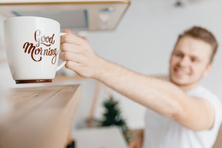 Man smile hand takes from shelf white ceramic mug for making breakfast coffee. Light sun, bright backgroundの写真素材