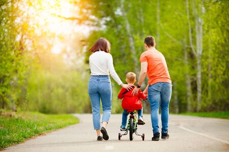 Dad and mom teaches little son to ride bike in park, have fun family, back view.の写真素材