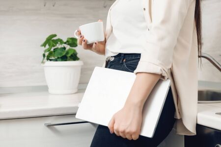 Young woman freelancer holds laptop and coffee in kitchen home. Concept work online quarantine.の写真素材