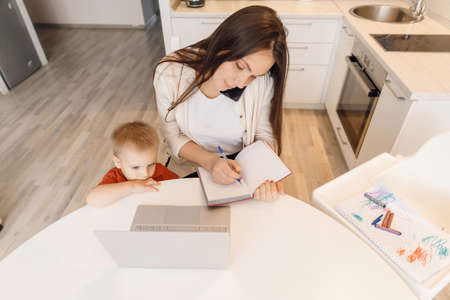 Woman on maternity leave working home online with laptop in kitchen with small child. Concept mom work while in quarantine isolation during Covid-19の写真素材