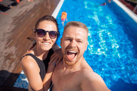 Couple of friends make selfie photo, happy caucasian beautiful young woman and man hug traveling on background hotel poolの写真素材