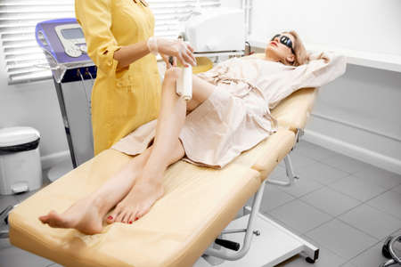 Beautician removes hair with medical laser from legs of beautiful woman in beauty salon, white backgroundの写真素材