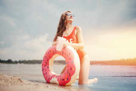 Happy woman resting on beach with inflatable ring with donut and cocktail, red swimsuit. Concept...