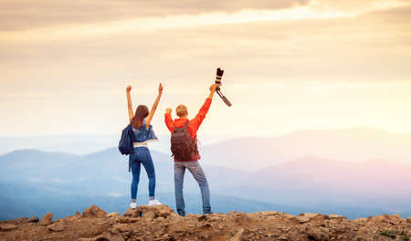Happy travelers couple conquered top of mountain, raises hands up with camera and takes photos of landscapes on tripの写真素材