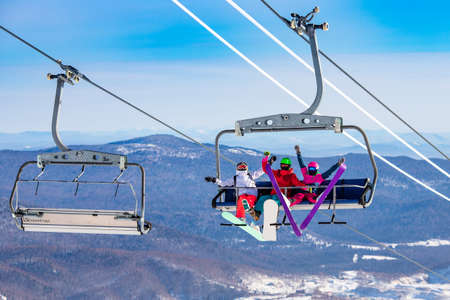 Fun team of friends skier and snowboarders up ski lift on blue sky backgroundの写真素材