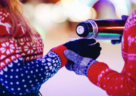 Loving couple drinks hot tea or coffee from thermal mugs on winter street in ice rink night illuminationの写真素材