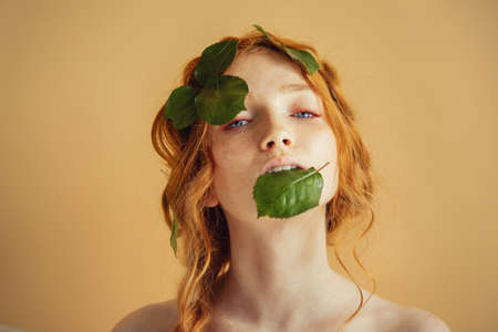 Beautiful young woman redhead blue eyes with green leaves on face. Nature organic cosmetics and makeupの写真素材