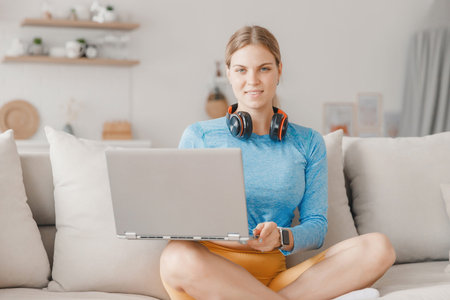 Young woman student in lotus position works online with laptop and headphones on couch against...