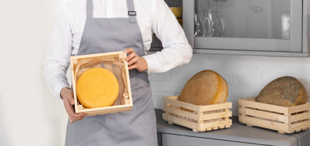 Banner farmer in gray apron holds homemade round of cheese in wooden box