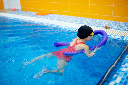 Child girl is engaged in swimming with trainer in pool, uses polyurethane stick to practice strokeの写真素材