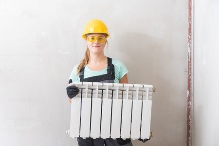Woman plumber holds white central heating radiator, in overalls and yellow hard hat against gray wallの写真素材