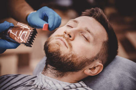 Close-up of barber with medical gloves shearing beard to man in barbershop. Haircut coronavirus pandemic protectionの写真素材