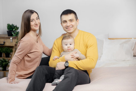 Happy parents smiling with small baby sitting on bed, looking into frame, light bedroom background