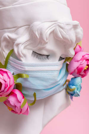 Sculpture of antique girl made of plaster in medical mask with flowers against pink background virus pandemicの写真素材