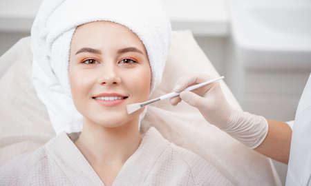 Doctor applies lifting cream or clearing acne skin with brush of young woman in beauty salonの写真素材