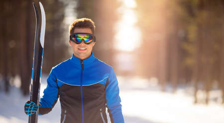 Portrait happy male athlete with cross country skis in hands and goggles, training in snowy forest. Healthy winter bannerの写真素材