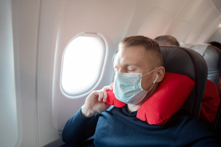 Male passenger of airplane listens to music and enjoys pillow for sleeping in chair. Concept travel with safe maskの写真素材