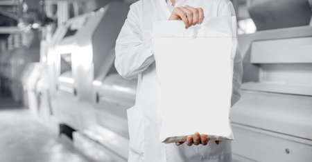 Operator holds bag of flour with products in background of automatic mill for wheat and cerealsの写真素材