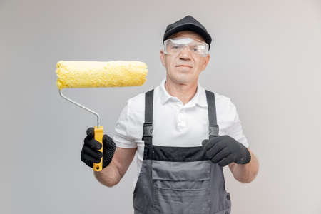 Old man builder painter in gloves and uniform holding painting roller repair apartment homeの写真素材