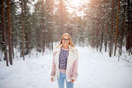 Smile young blonde woman in glasses, pink jacket in winter clothes run snowy forest sunlightの写真素材