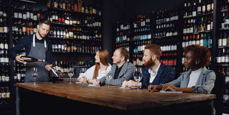 Group of European friends and African woman at wine tasting master class in restaurant, sommelier pours drink into decanterの写真素材