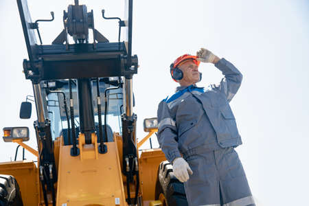 Man in helmet in front of bulldozer or excavator at construction site. Concept of interaction of technology and industrial workerの写真素材