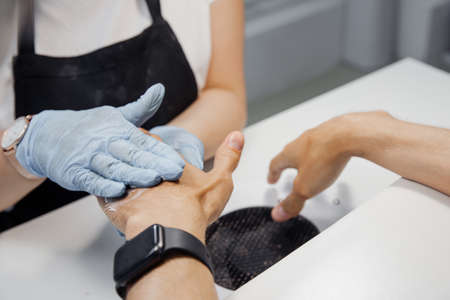 Young man gets professional manicure care for nail in spa salon, master applies hand creamの写真素材