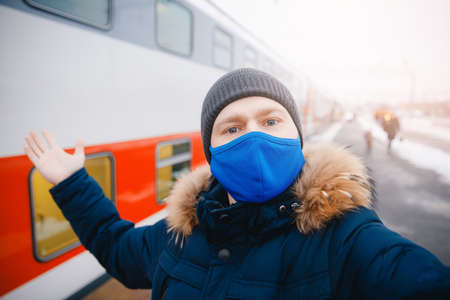 Winter train journey, happy young man wearing protective mask on railway platform arrives at station and make selfie photoの写真素材