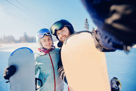Happy lovers couple man and young women snowboarders make selfie photo on background of ski resort, sun lightの写真素材