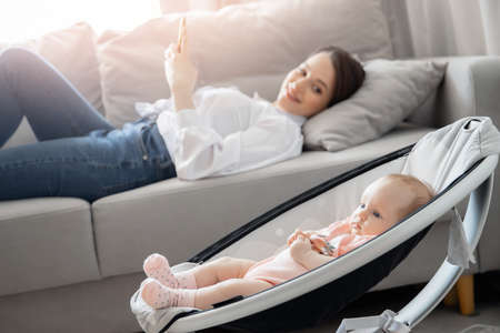 Young woman mom works internet with phone on home office while baby sleeps in automatic cradle, sun lightの写真素材