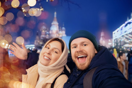 Happy friends man and woman making selfie photo and walking in night city Christmas market, bokeh lights. Winter vacations in Red square Moscowの写真素材