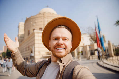 Young archaeologist man in hat takes selfie photo against background of ancient buildings. Concept archeological sit old cityの写真素材