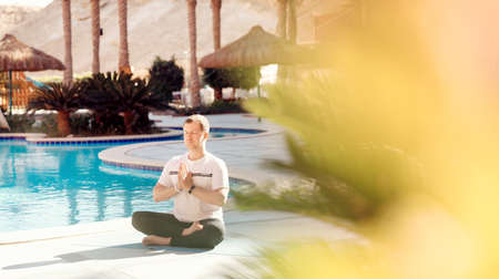 Meditation yoga caucasian instructor man conducts classes by hotel pool on background palm trees and sunlightの写真素材