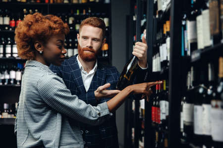 Lover multicultural couple talking and choosing wine in shop. Man and African girl are buying alcoholの写真素材