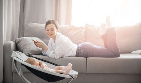 Young woman mom works internet with phone on home office while baby sleeps in automatic cradle, sun lightの写真素材