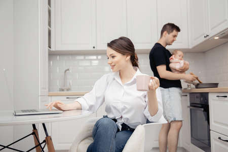 Man was laid off work due to coronavirus, young woman mother working online at computer. Father is babysitting and preparing food in kitchenの写真素材