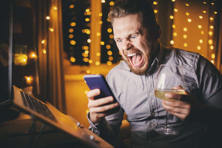Bearded man home screaming and angry with phone and laptop. Concept anger issues and stressの写真素材